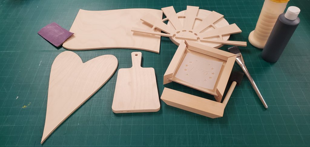 A picture of some wood craft supplies ready to be decorated. Includes a stencil, paint brushes, some black paint, and wood glue.